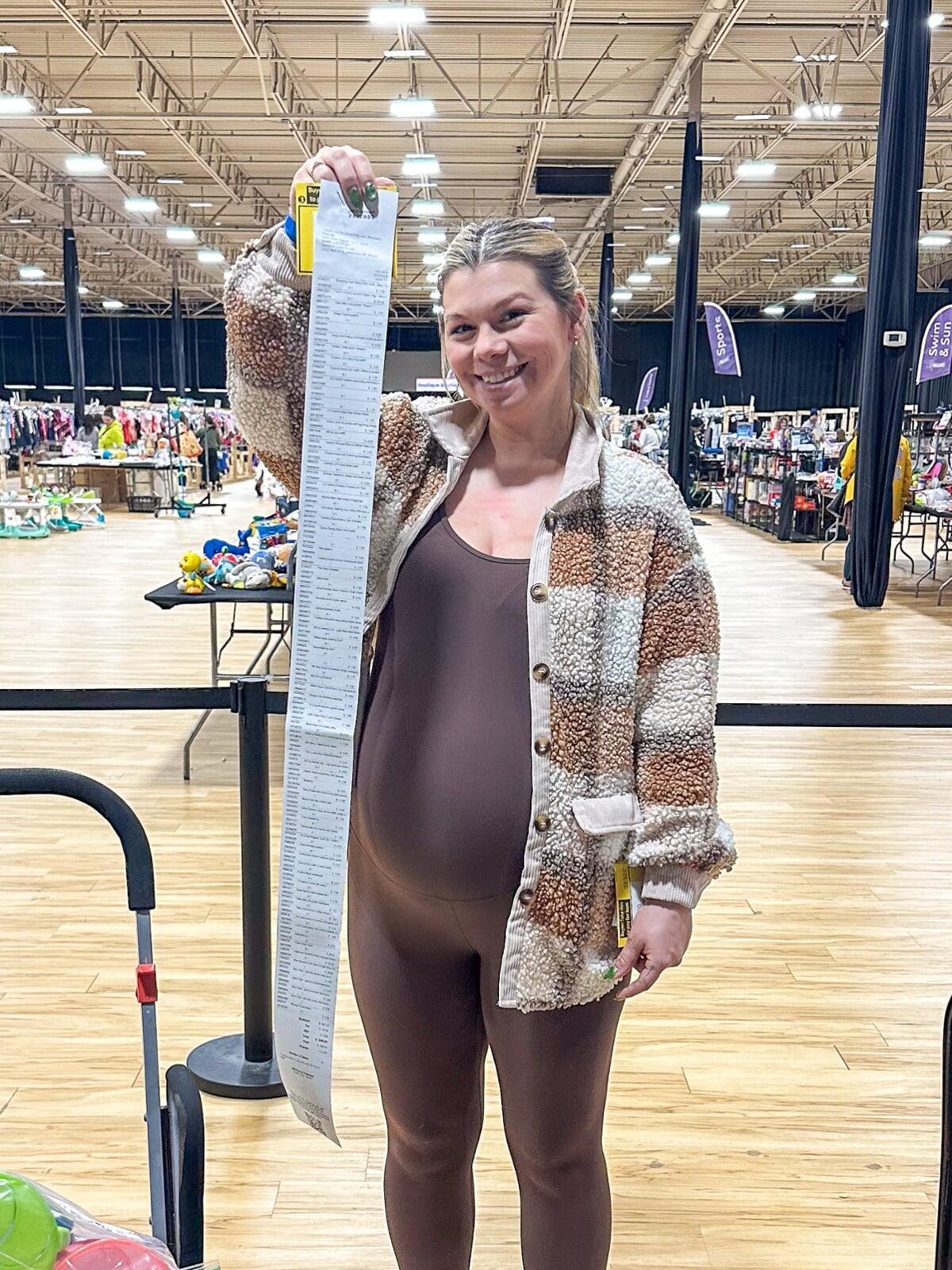 A Mom holds up her JBF shopping bag with her right hand while resting her left hand on her stroller with toddler daughter inside.
